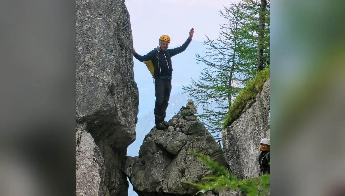 Ein Bergsteiger steht mit erhobenen Armen auf einem Felsblock im Fensterl, hinten Wald und Bergpanorama. | © DAV Markt Schwaben · Foto: Erwin Matzinger
