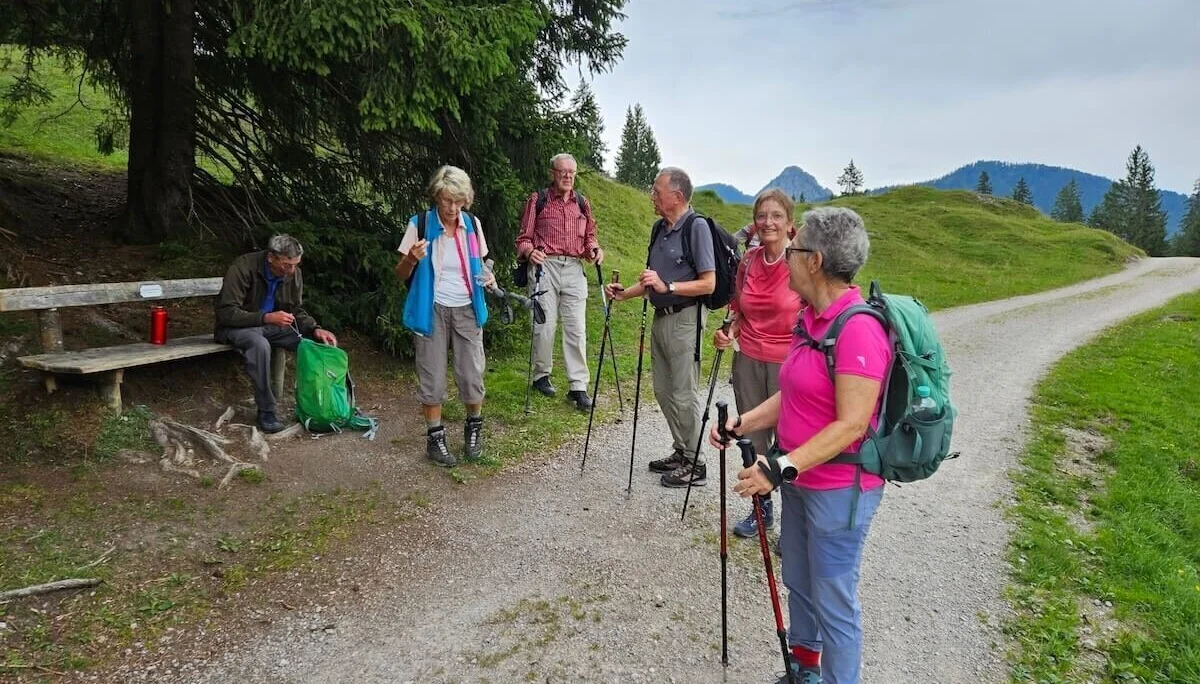 Fünf Wandernde stehen auf einem Bergpfad, eine Person sitzt auf einer Bank. Im Hintergrund: Wiesen, Wald und Bergpanorama. | © DAV Markt Schwaben | Foto Hubert Inhofer