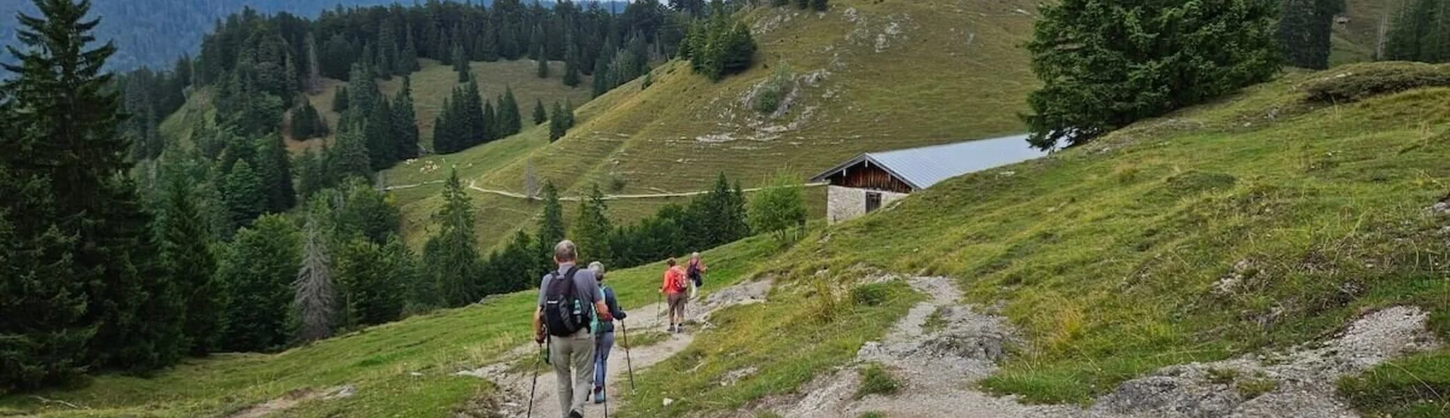 Drei Wandernde mit Rucksäcken und Stöcken auf einem Bergpfad, im Hintergrund eine Holzhütte und bewaldete Hänge. | © DAV Markt Schwaben | Foto Hubert Inhofer