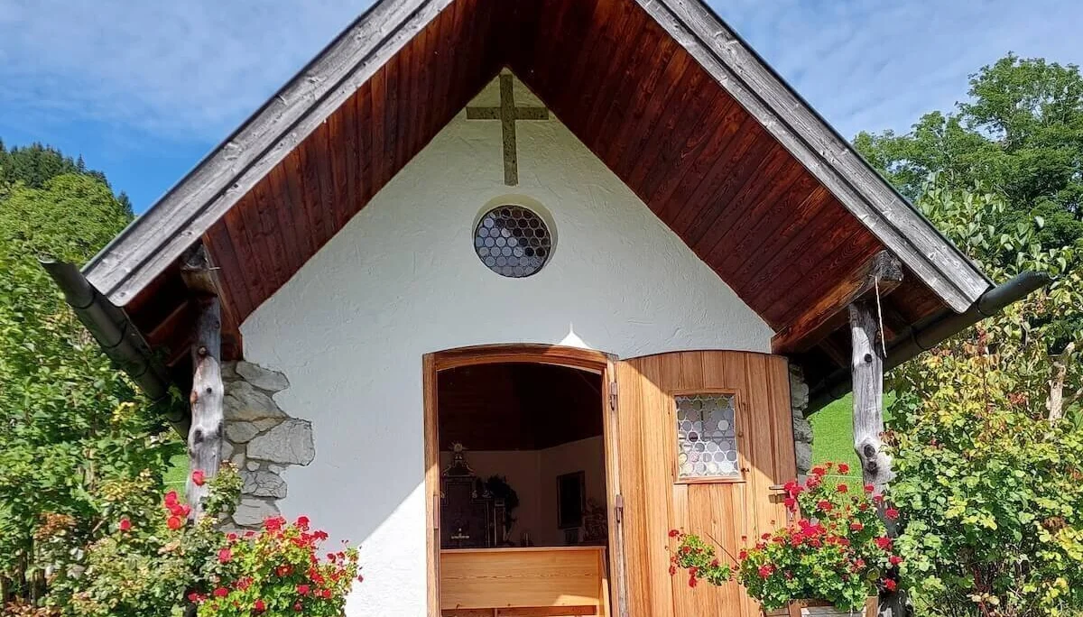 Weiße Kapelle mit dunklem Holzdach und roten Geranien, umgeben von Bäumen unter leicht bewölktem Himmel. | © DAV Markt Schwaben | Foto Hubert Inhofer