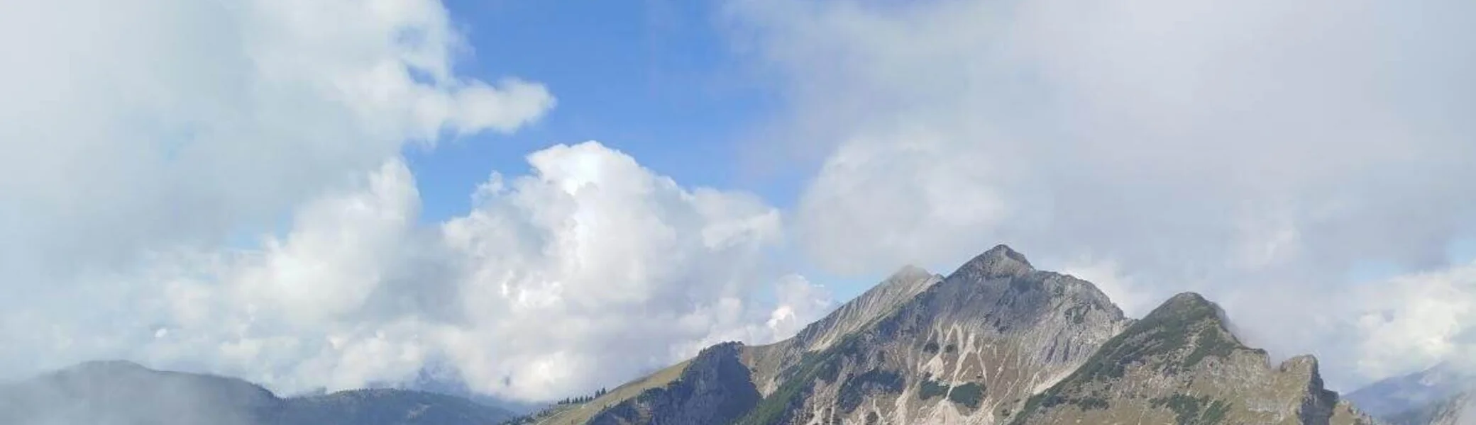 Drei Wandernde stehen vor rustikaler Almhütte aus Holz und Stein; hügelige Landschaft und Wolken im Hintergrund. | © DAV Markt Schwaben | Foto Helga Peters