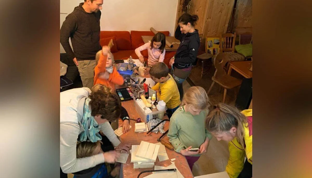 Kinder und Erwachsene gestalten am Basteltisch farbenfrohe Holzobjekte; im Hintergrund ein Holzspielhaus. | © DAV Markt Schwaben | Foto: Helena De Monte