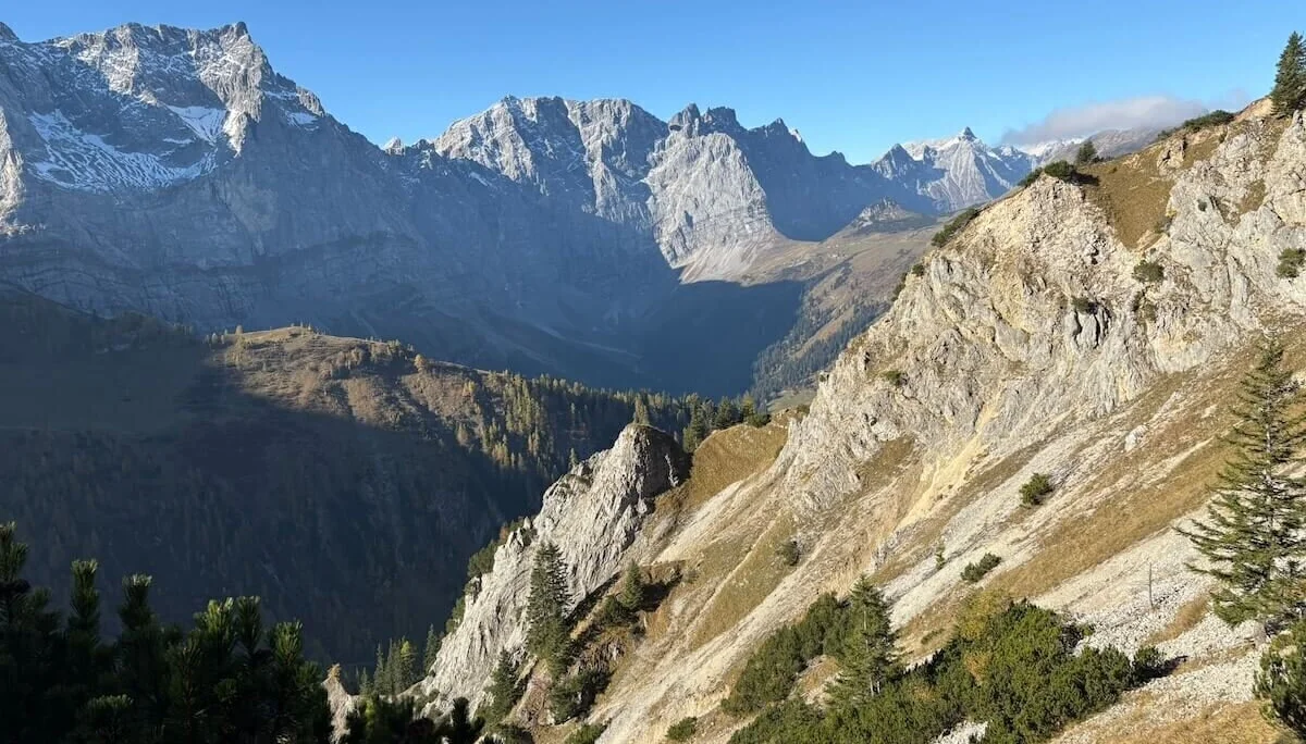 Im Vordergrund felsiges Gelände mit Nadelbäumen, dahinter schneebedeckte Berggipfel und bewaldete Täler bei Sonnenschein. | © DAV Markt Schwaben · Foto: Markus Zimmermann