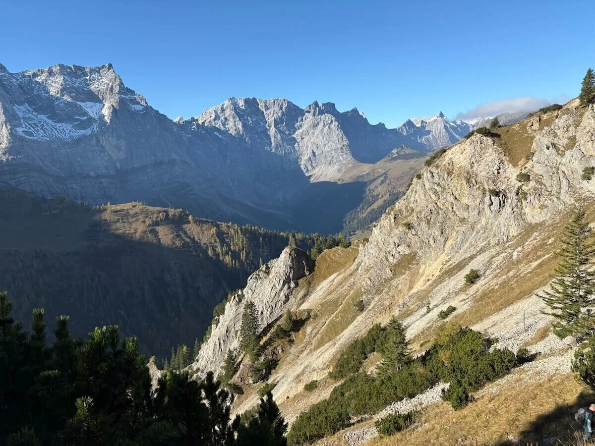 Im Vordergrund felsiges Gelände mit Nadelbäumen, dahinter schneebedeckte Berggipfel und bewaldete Täler bei Sonnenschein. | © DAV Markt Schwaben · Foto: Markus Zimmermann