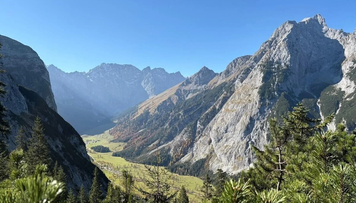 Grüne Nadelbäume im Vordergrund rahmen ein tiefes Tal mit Wiesen und Felsen; dahinter steile Bergflanken bei Sonnenschein. | © DAV Markt Schwaben · Foto: Markus Zimmermann