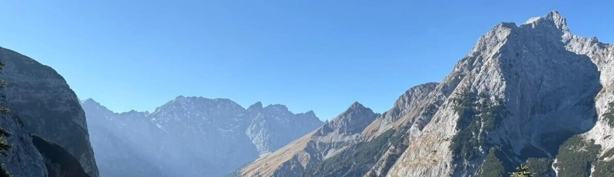 Grüne Nadelbäume im Vordergrund rahmen ein tiefes Tal mit Wiesen und Felsen; dahinter steile Bergflanken bei Sonnenschein. | © DAV Markt Schwaben · Foto: Markus Zimmermann