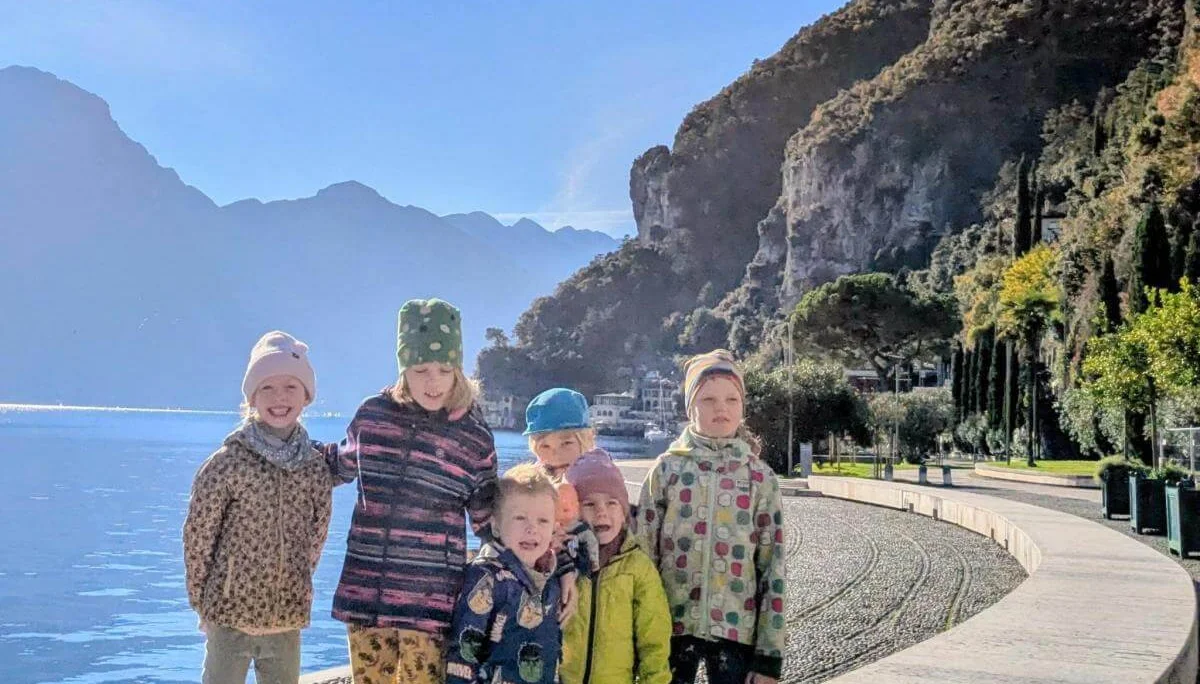 Mehrere Erwachsene und Kinder sitzen auf Matten und Baumstämmen in einem herbstlichen Waldstück. Ein Kind steht in der Mitte mit erhobenen Armen. Alle tragen warme Kleidung, im Hintergrund sind bunte Blätter, ein Zaun und Berge zu sehen. | © DAV Markt Schwaben · Foto: Silke Titze