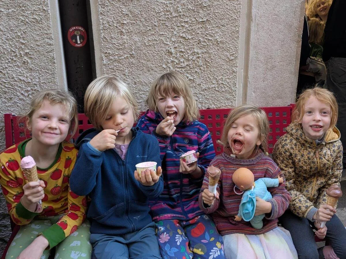 Drei Kinder in Kletterausrüstung stehen auf einer Wiese neben einer schwarzen Ziege. Ein Kind hält einen Ast, vermutlich zum Füttern. Im Hintergrund klettern weitere Personen an einem Felsen, umgeben von Bäumen und Sonnenlicht. | © DAV Markt Schwaben · Foto: Silke Titze