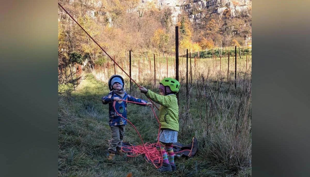 Ein Kind mit rotem Helm und Kletterausrüstung steigt eine steile Felswand hinauf. Dahinter klettert eine weitere Person mit blauer Ausrüstung. Beide sind gesichert und konzentriert in Bewegung. Die Szene spielt sich an einem sonnigen Felsen in der Natur ab. | © DAV Markt Schwaben · Foto: Silke Titze