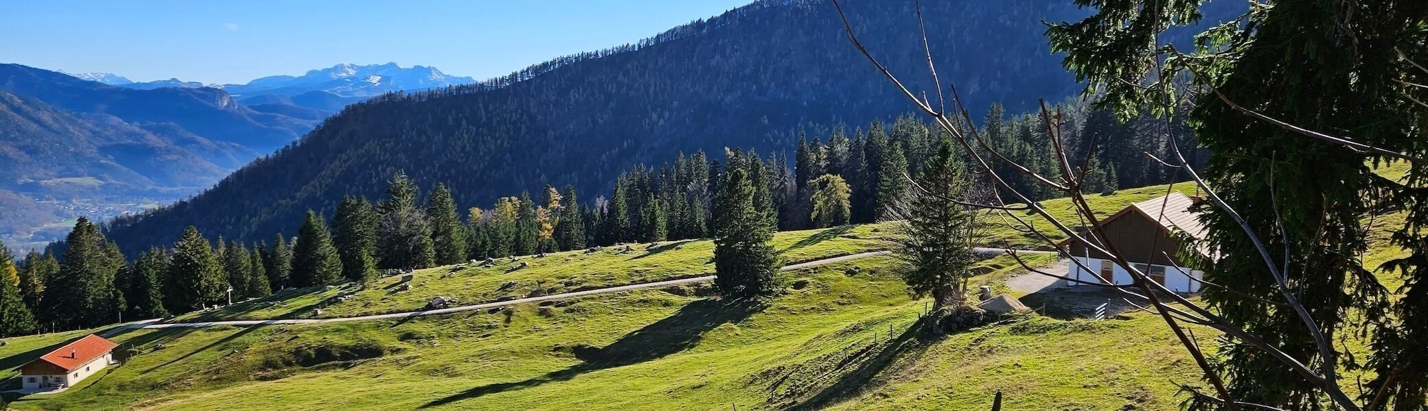 Zwei Hütten auf grüner Wiese im Vordergrund, dahinter Wald, Weg und schneebedeckte Berge unter blauem Himmel. | © DAV Markt Schwaben · Foto: Hubert Inhofer