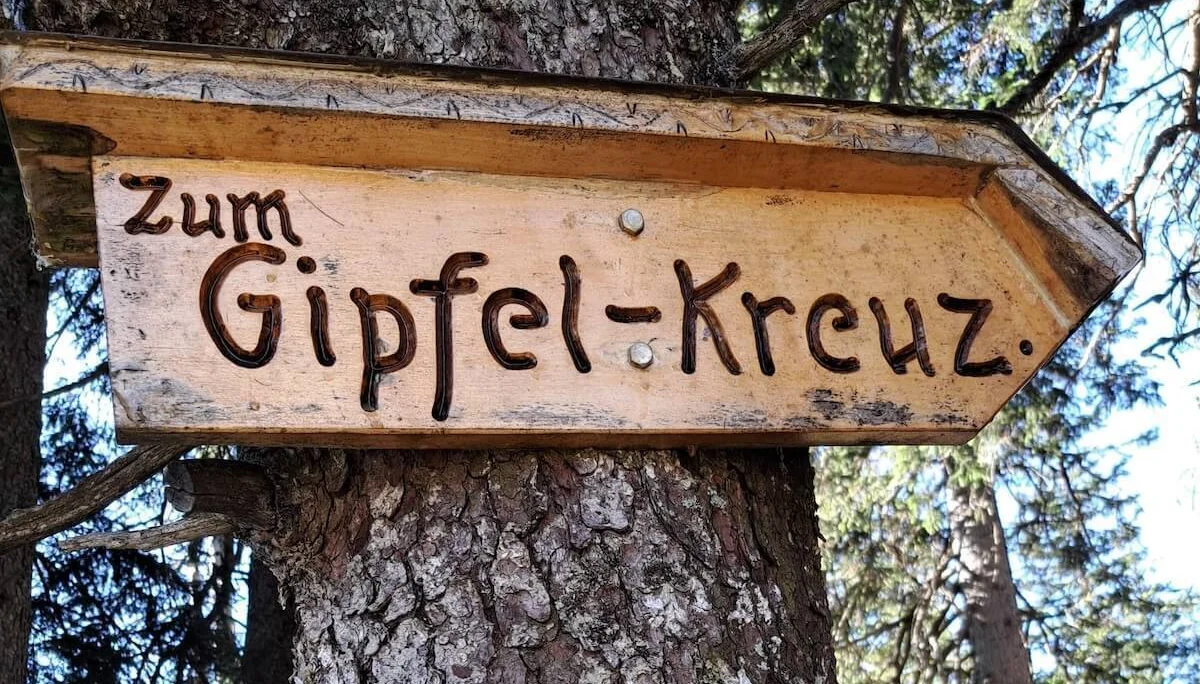 Ein hölzerner Richtungspfeil mit der Aufschrift „Zum Gipfel-Kreuz“ ist an einem Baum befestigt. Der Wegweiser zeigt nach rechts und fügt sich rustikal in die bewaldete Umgebung ein. Er markiert den Pfad zum Gipfel und vermittelt Orientierung und Vorfreude auf das Ziel. | © DAV Markt Schwaben | Foto: Tanja Döhler