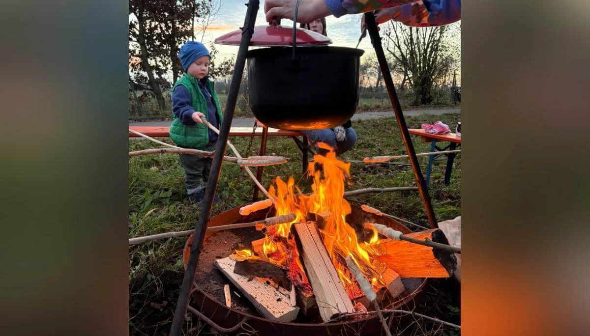 Ein Kind mit warmer Kleidung steht neben einem Lagerfeuer und hält einen Stock ins Feuer. Ein Erwachsener hebt den Deckel eines schwarzen Topfs, der über dem Feuer hängt. Rundherum liegen Holzscheite, im Hintergrund sind Bäume und Sitzbänke sichtbar. | © DAV Markt Schwaben · Foto: Lara Wolff