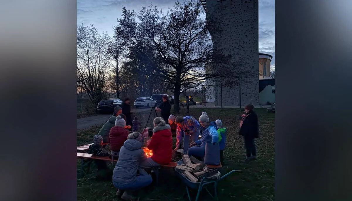 Mehrere Personen sitzen bei Dämmerung um ein Lagerfeuer, warm gekleidet mit Jacken und Mützen. Sie unterhalten sich oder schauen ins Feuer. Im Hintergrund stehen ein Kletterturm, ein rundes Gebäude und geparkte Autos. Die Bäume sind kahl, es wirkt herbstlich. | © DAV Markt Schwaben · Foto: Lara Wolff