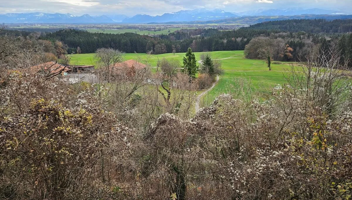 Vorne Sträucher und Bauernhof, mittig Wiesen mit Weg, hinten Wald und schneebedeckte Berge unter Wolken. | © DAV Markt Schwaben · Foto: Hubert Inhofer