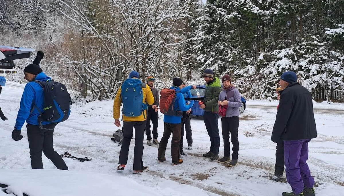 Winterwandergruppe mit Rucksäcken und Stöcken steht auf verschneitem Waldboden, Bäume und Fahrzeug im Hintergrund. | © DAV Markt Schwaben · Foto Tanja Döhler