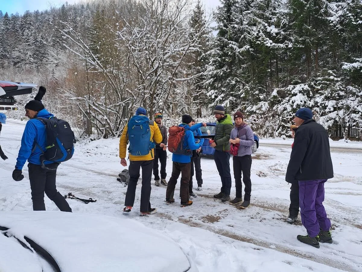 Winterwandergruppe mit Rucksäcken und Stöcken steht auf verschneitem Waldboden, Bäume und Fahrzeug im Hintergrund. | © DAV Markt Schwaben · Foto Tanja Döhler