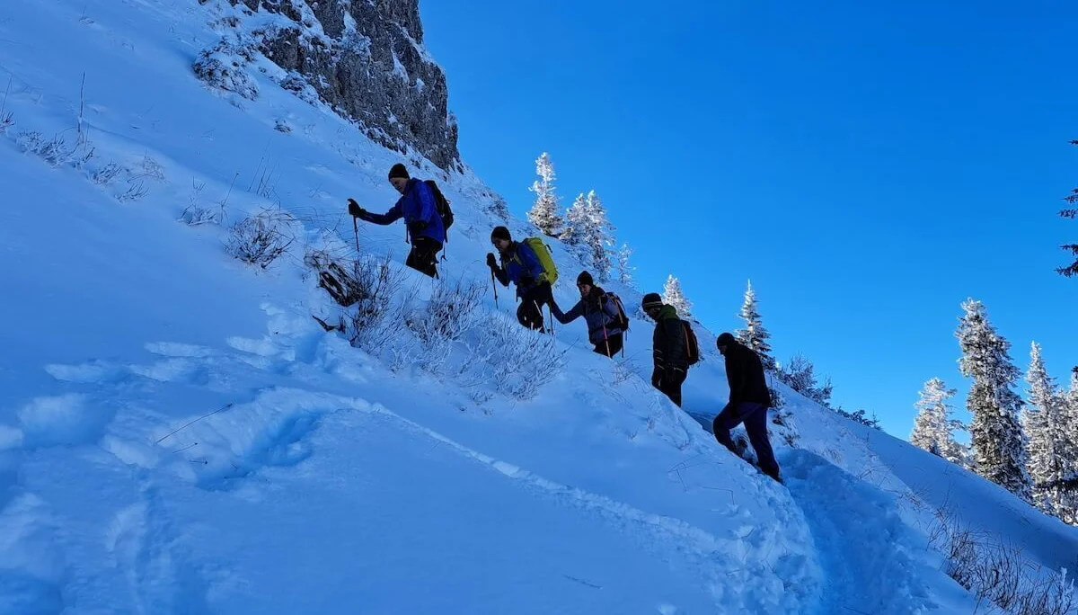 Sechs Personen steigen mit Winterausrüstung und Stöcken einen verschneiten Bergpfad hinauf, blauer Himmel im Hintergrund. | © DAV Markt Schwaben · Foto Tanja Döhler