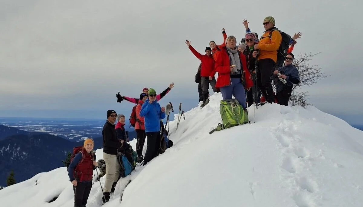Eine Gruppe von Winterwandernden steht auf einem verschneiten Gipfel und posiert mit erhobenen Armen. Im Hintergrund sind ferne Bergketten und ein wolkenverhangener Himmel zu sehen. | © DAV Markt Schwaben · Foto Alfred Quiel