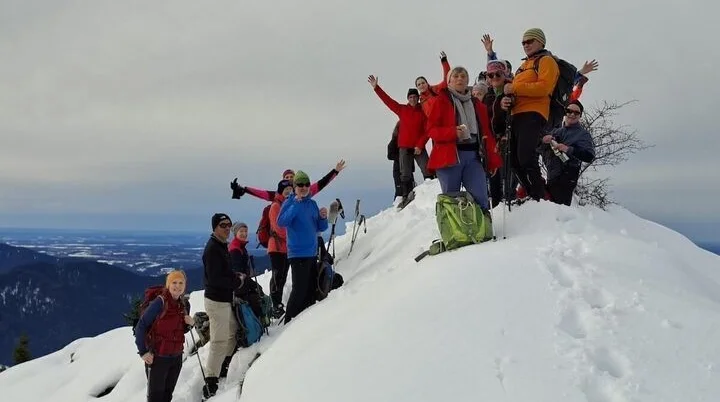 Eine Gruppe von Winterwandernden steht auf einem verschneiten Gipfel und posiert mit erhobenen Armen. Im Hintergrund sind ferne Bergketten und ein wolkenverhangener Himmel zu sehen. | © DAV Markt Schwaben · Foto Alfred Quiel