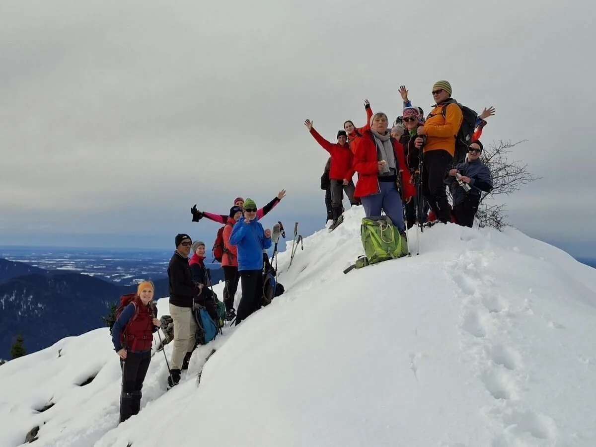 Eine Gruppe von Winterwandernden steht auf einem verschneiten Gipfel und posiert mit erhobenen Armen. Im Hintergrund sind ferne Bergketten und ein wolkenverhangener Himmel zu sehen. | © DAV Markt Schwaben · Foto Alfred Quiel