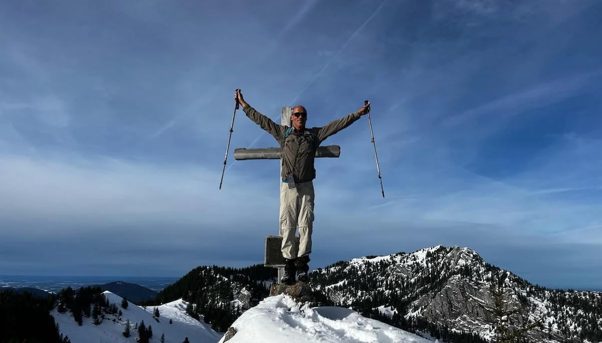 Ein Bergsteiger steht mit erhobenen Wanderstöcken neben einem Gipfelkreuz auf einem verschneiten Berg. Im Hintergrund erstreckt sich ein Panorama aus schneebedeckten Gipfeln unter blauem Himmel mit Wolken. | © DAV Markt Schwaben · Foto Markus Sellmeier