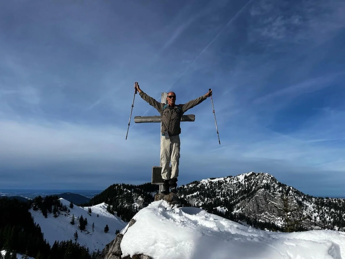 Ein Bergsteiger steht mit erhobenen Wanderstöcken neben einem Gipfelkreuz auf einem verschneiten Berg. Im Hintergrund erstreckt sich ein Panorama aus schneebedeckten Gipfeln unter blauem Himmel mit Wolken. | © DAV Markt Schwaben · Foto Markus Sellmeier