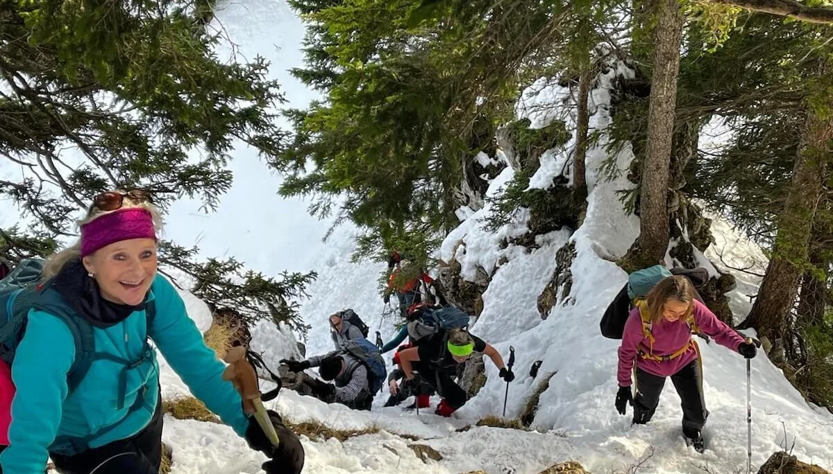 Eine Wanderin mit Winterausrüstung steigt einen steilen, verschneiten Bergpfad hinauf, umgeben von Nadelbäumen. Die Szene zeigt die anspruchsvolle Passage in alpinem Gelände. | © DAV Markt Schwaben · Foto Markus Sellmeier