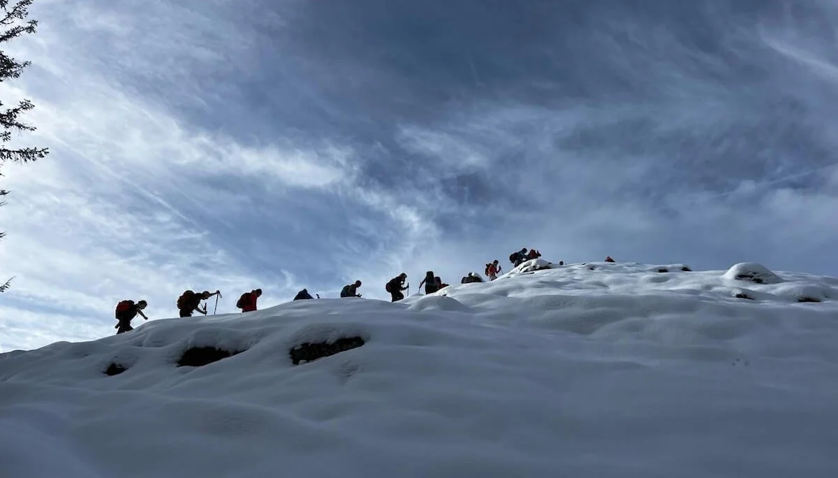Eine Gruppe von Wandernden steigt mit Rucksäcken und Stöcken einen verschneiten Hang hinauf. Der Himmel ist teils bewölkt, die Personen sind als Silhouetten vor der dramatischen Wolkenkulisse sichtbar. | © DAV Markt Schwaben · Foto Markus Sellmeier