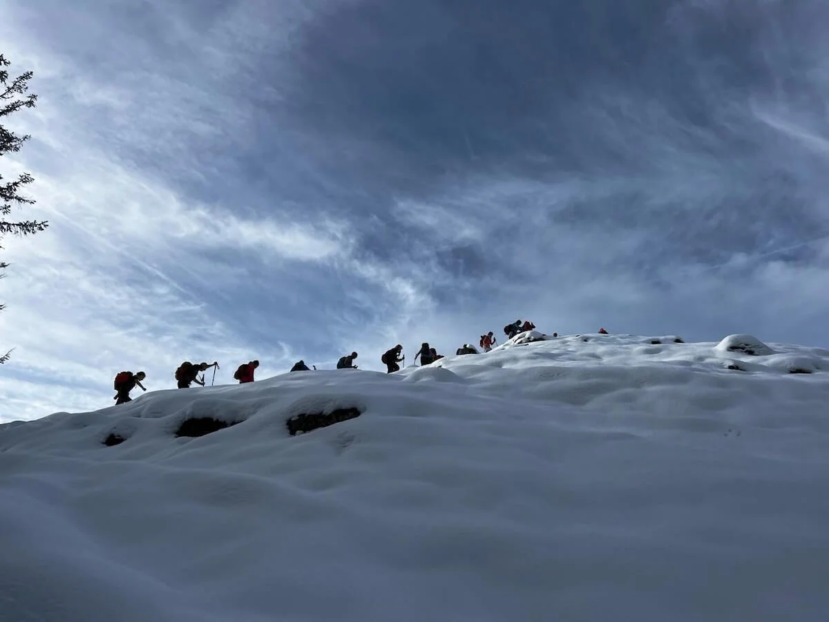 Eine Gruppe von Wandernden steigt mit Rucksäcken und Stöcken einen verschneiten Hang hinauf. Der Himmel ist teils bewölkt, die Personen sind als Silhouetten vor der dramatischen Wolkenkulisse sichtbar. | © DAV Markt Schwaben · Foto Markus Sellmeier