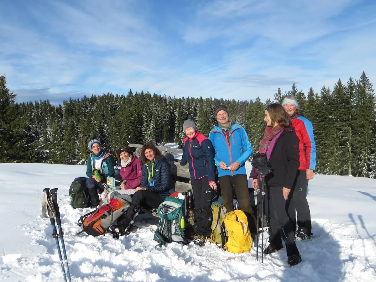 Sieben Wandernde pausieren an einer Holzbank im Schnee; im Hintergrund verschneite Tannen und blauer Himmel mit Wolken. | © DAV Markt Schwaben | Foto: Gerlinde Hübl