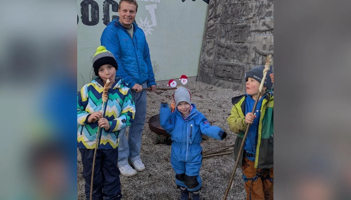 Vier Kinder mit Stöcken vor bemalter Wand und Feuerstelle; Winterkleidung, Erwachsene im Hintergrund, Kiesplatz. | © DAV Markt Schwaben · Familiengruppe