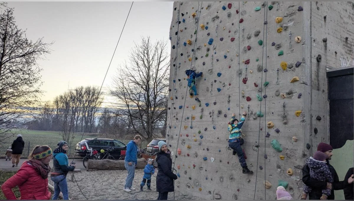 Zwei Kinder klettern gesichert an der bunten Kletterwand; Erwachsene stehen daneben, Bäume im Hintergrund. | © DAV Markt Schwaben · Familiengruppe