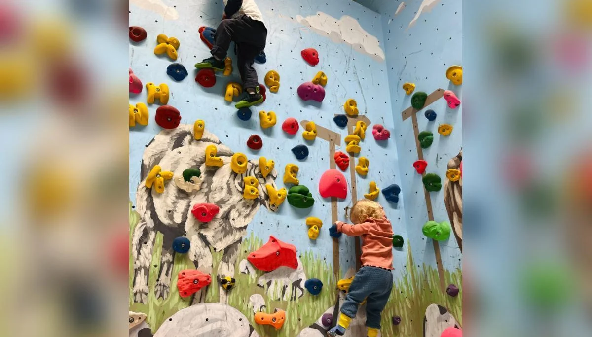 Zwei Kinder klettern an bunten Griffen vor einer Wand mit Tiermotiven; Schaf und Kuh im Hintergrund, Fokus auf Bewegung. | © DAV Markt Schwaben | Foto: Lara Wolff
