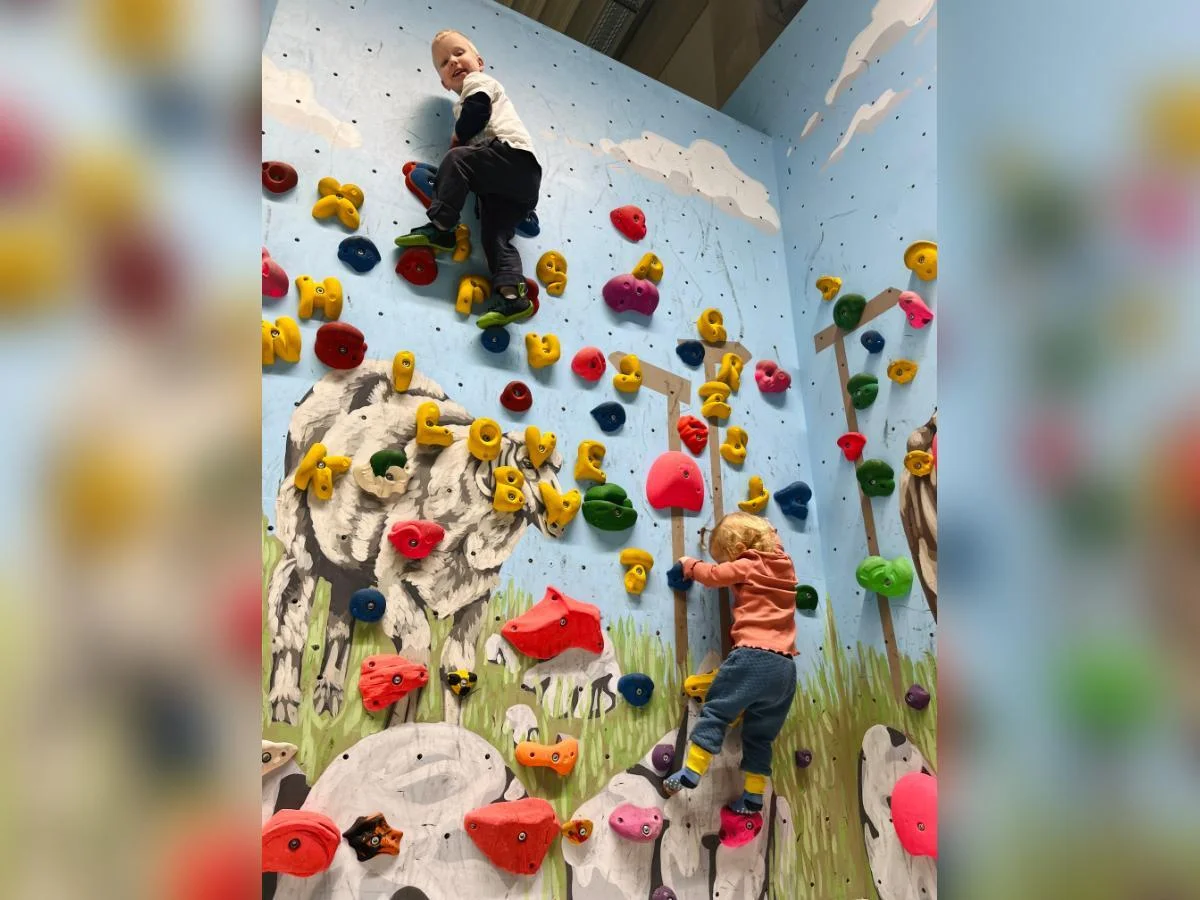 Zwei Kinder klettern an bunten Griffen vor einer Wand mit Tiermotiven; Schaf und Kuh im Hintergrund, Fokus auf Bewegung. | © DAV Markt Schwaben | Foto: Lara Wolff
