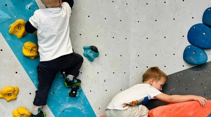 Zwei Kinder klettern konzentriert an bunten Bouldergriffen; geometrisch gestaltete Wand im Hintergrund zeigt Routenvielfalt. | © DAV Markt Schwaben | Foto: Lara Wolff