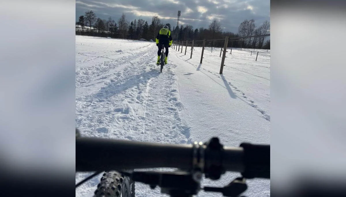 Lenker im Vordergrund, ein Radfahrer voraus auf verschneitem Weg; Zaunpfähle und Bäume rahmen die winterliche Szene. | © DAV Markt Schwaben | Foto: Miron Thoms