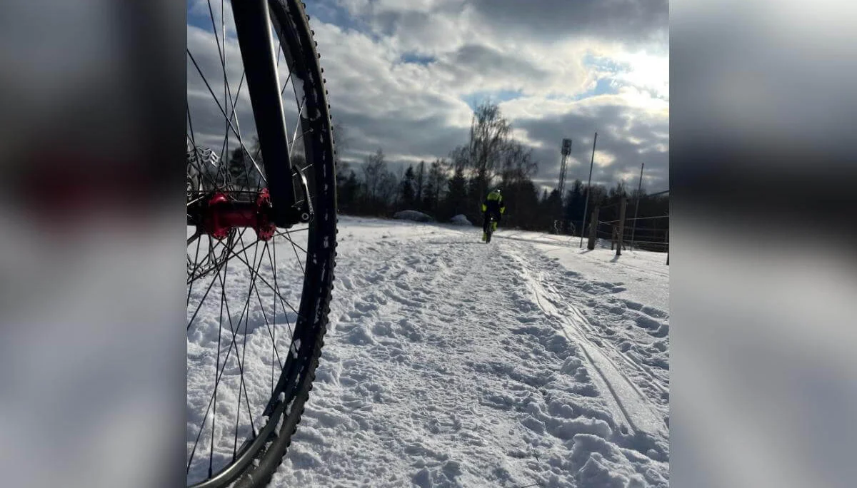 Nahaufnahme eines Fahrradreifens links im Vordergrund, dahinter fährt eine Person auf verschneitem Weg Richtung Horizont. | © DAV Markt Schwaben | Foto: Miron Thoms