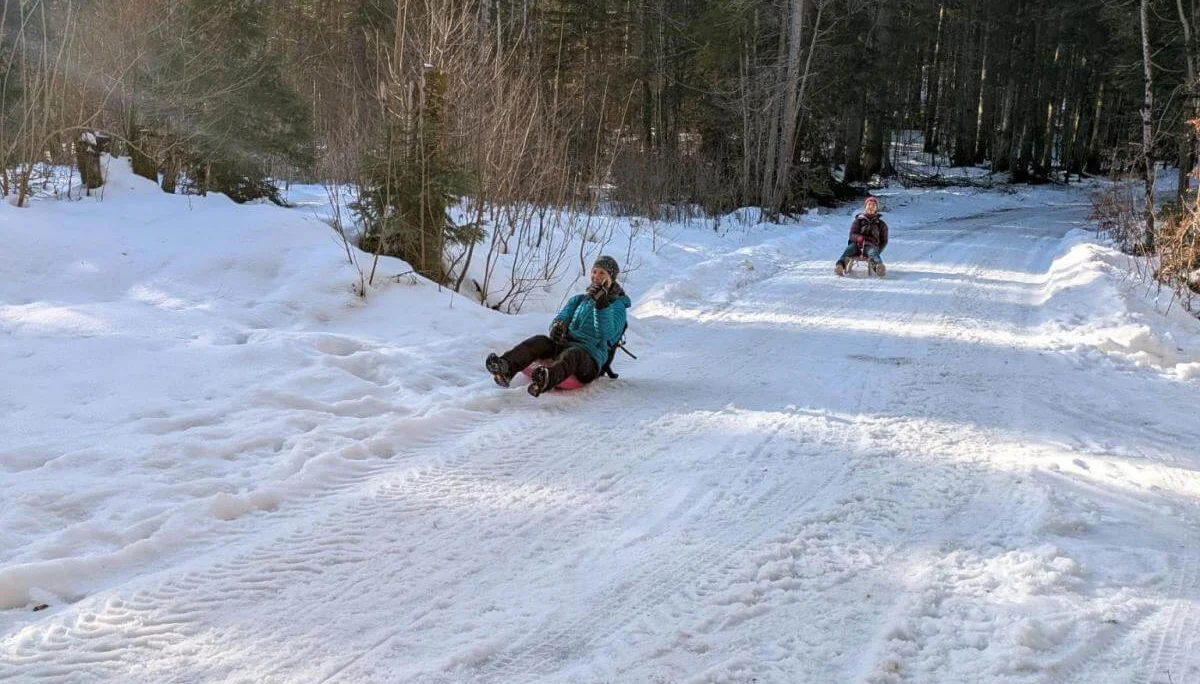 Zwei Erwachsene rodeln auf bunten Schlitten einen verschneiten Waldweg hinunter, Sonnenlicht fällt durch die Bäume. | © DAV Markt Schwaben | Foto: Silke Titze