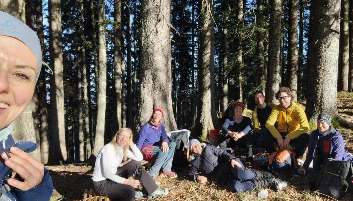 Acht Erwachsene sitzen entspannt im Wald zwischen Rucksäcken und Sonnenstrahlen, eine Person macht ein Selfie. | © DAV Markt Schwaben | Foto: Silke Titze