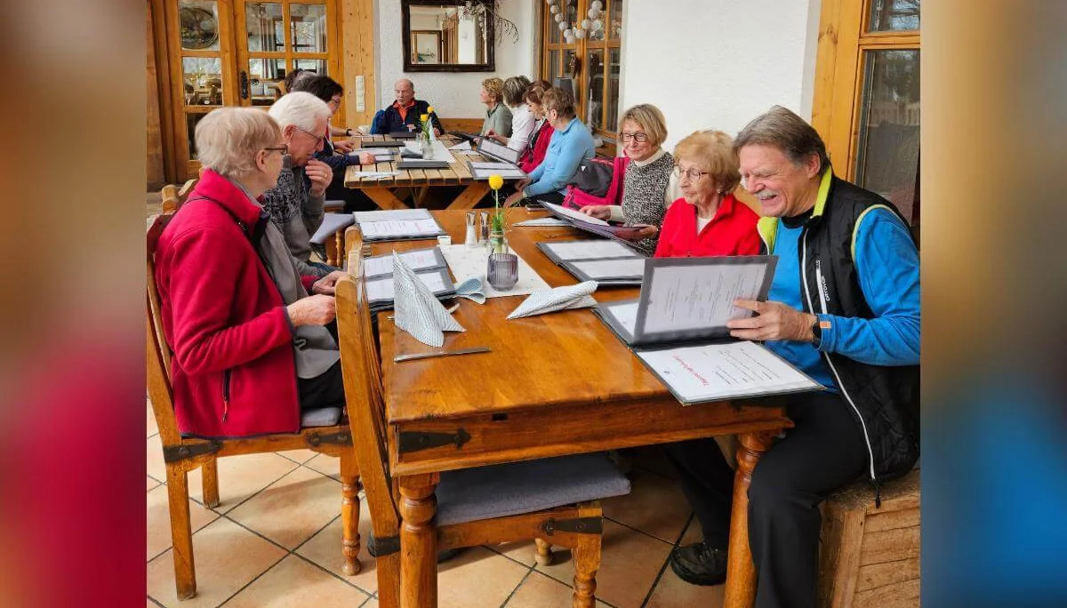 Seniorinnen und Senioren sitzen in einer warmen Stube an zwei Holztischen und lesen Speisekarten; durch große Fenster fällt Tageslicht. | © DAV Markt Schwaben | Foto: Hubert Inhofer