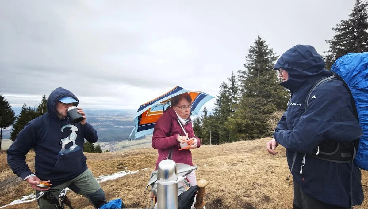 Drei Wandernde machen an einem Holztisch Pause; sie trinken und essen, im Hintergrund liegt ein weites Tal unter bewölktem Himmel. | © DAV Markt Schwaben | Foto: Erwin Matzinger