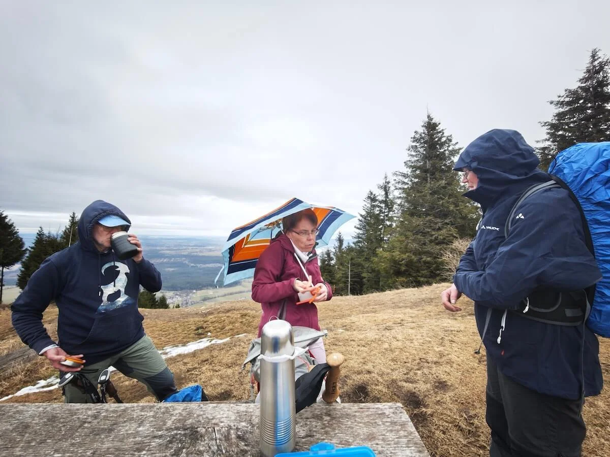 Drei Wandernde machen an einem Holztisch Pause; sie trinken und essen, im Hintergrund liegt ein weites Tal unter bewölktem Himmel. | © DAV Markt Schwaben | Foto: Erwin Matzinger