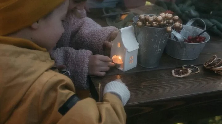 Zwei Kinder betrachten neugierig ein kleines, beleuchtetes Häuschen auf einem Holztisch im Freien. Der Tisch ist herbstlich dekoriert mit Eicheln, roten Beeren und getrockneten Orangenscheiben. | © Free to use photo from Pexels