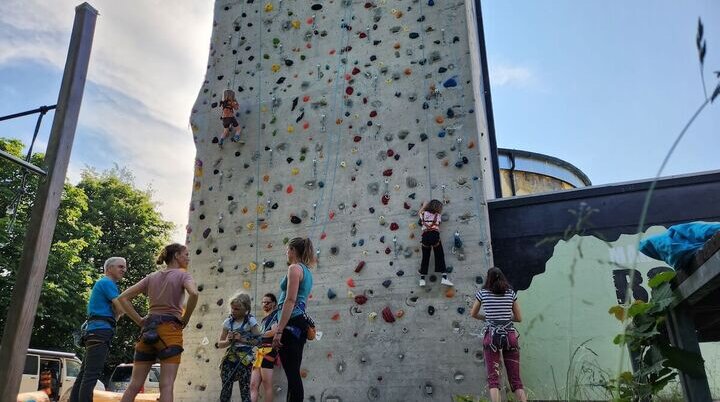 Mehrere Personen klettern eine Outdoor-Kletterwand mit bunten Griffen hinauf. Zwei Kinder sind aktiv am Klettern, während andere daneben stehen, beobachten oder warten. | © DAV Markt Schwaben