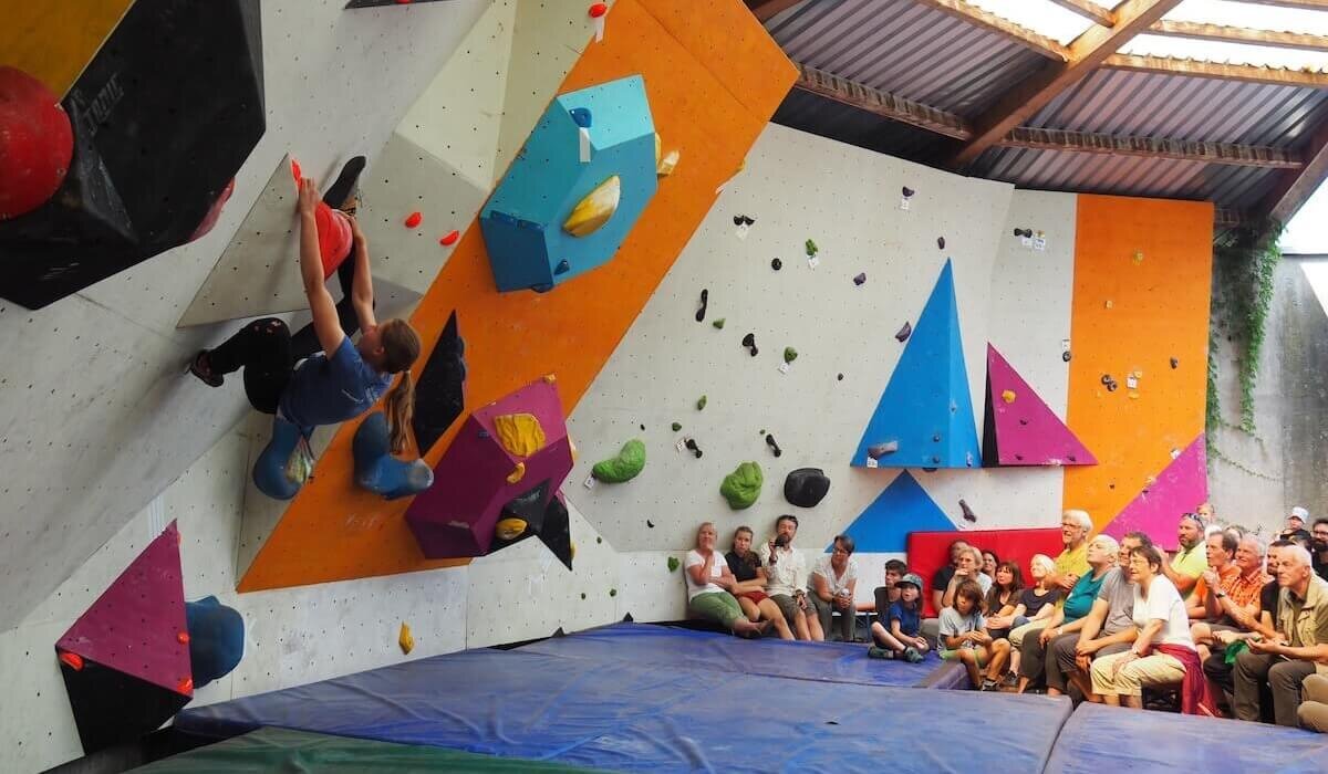 Boulder-Cup 2023 | © DAV Markt Schwaben | Foto Elke Kressirer Ein Kletterer erklimmt eine Boulderwand, beobachtet von einer Gruppe Zuschauer in einer lichtdurchfluteten Kletterhalle während des Wettkampfs. | © DAV Markt Schwaben | Foto Elke Kressirer
