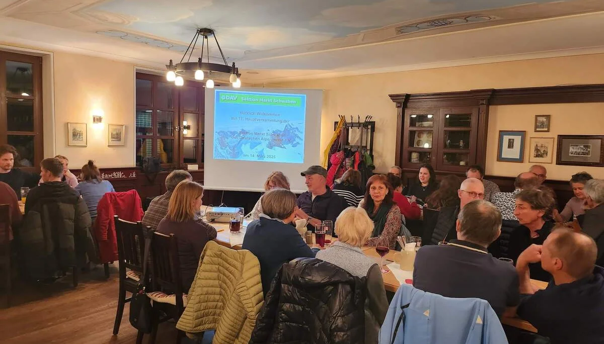 Mitglieder der DAV Sektion Markt Schwaben sitzen in einem Raum und schauen eine Präsentation auf einer Leinwand mit 'Herzlich Willkommen zur 17. Hauptversammlung DAV Sektion Markt Schwaben'. | © DAV Markt Schwaben | Foto Anna Thoms