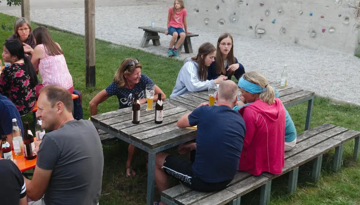 Menschen sitzen an Bierbänken im Freien, unterhalten sich und genießen Getränke, während im Hintergrund der Kletterturm zu sehen ist. | © DAV Markt Schwaben/Thomas Schäffler
