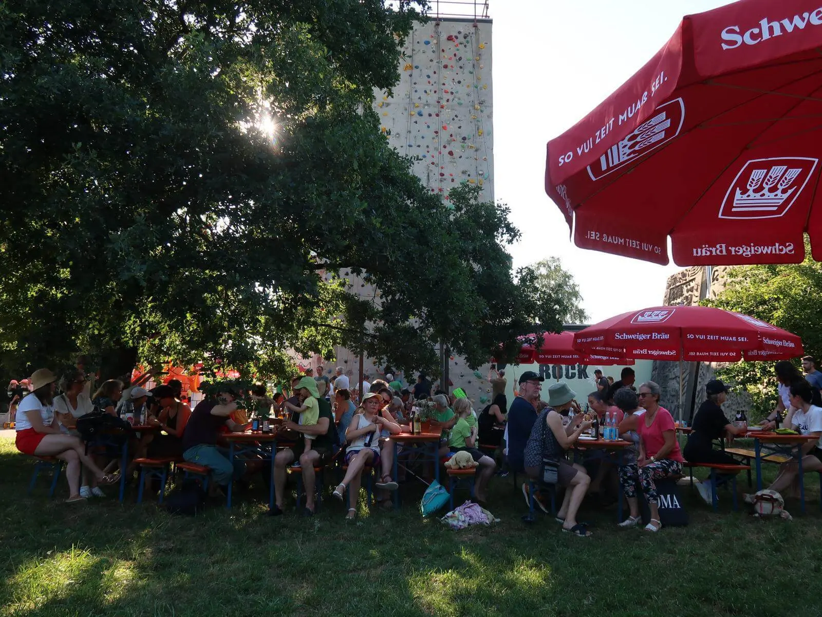 Menschen sitzen unter roten Sonnenschirmen, im Hintergrund steht die DAV‑Kletterwand. | © DAV Markt Schwaben/Susanne Mayerl