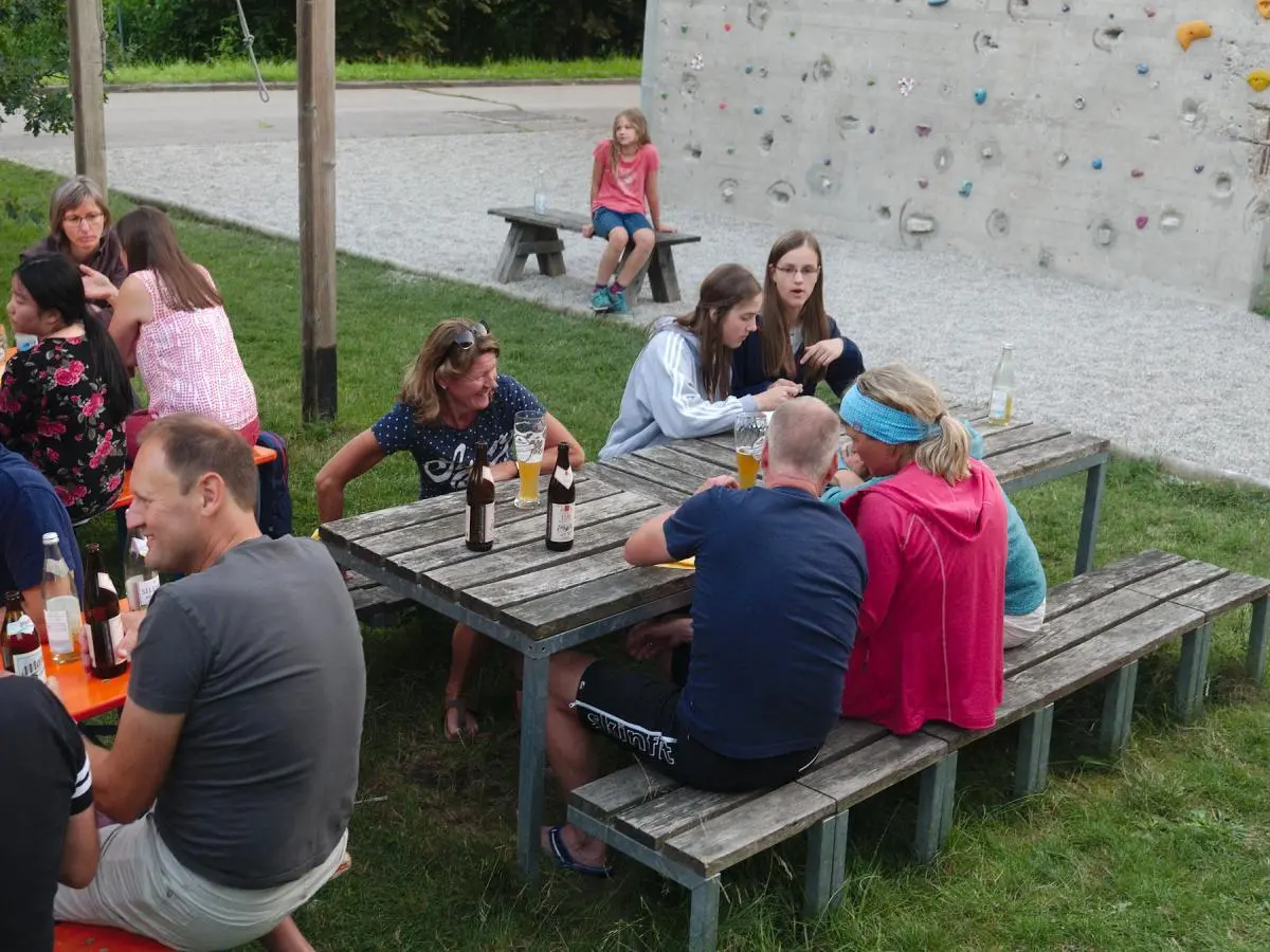 Menschen sitzen an Bierbänken im Freien, unterhalten sich und genießen Getränke, während im Hintergrund der Kletterturm zu sehen ist. | © DAV Markt Schwaben/Thomas Schäffler