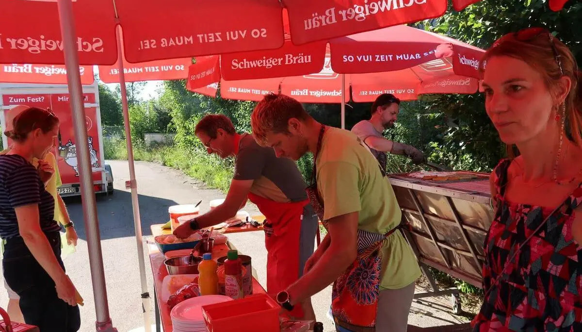 Team bereitet Essen am Grillstand unter roten Sonnenschirmen beim Sommerfest zu. | © DAV Markt Schwaben/Susanne Mayerl